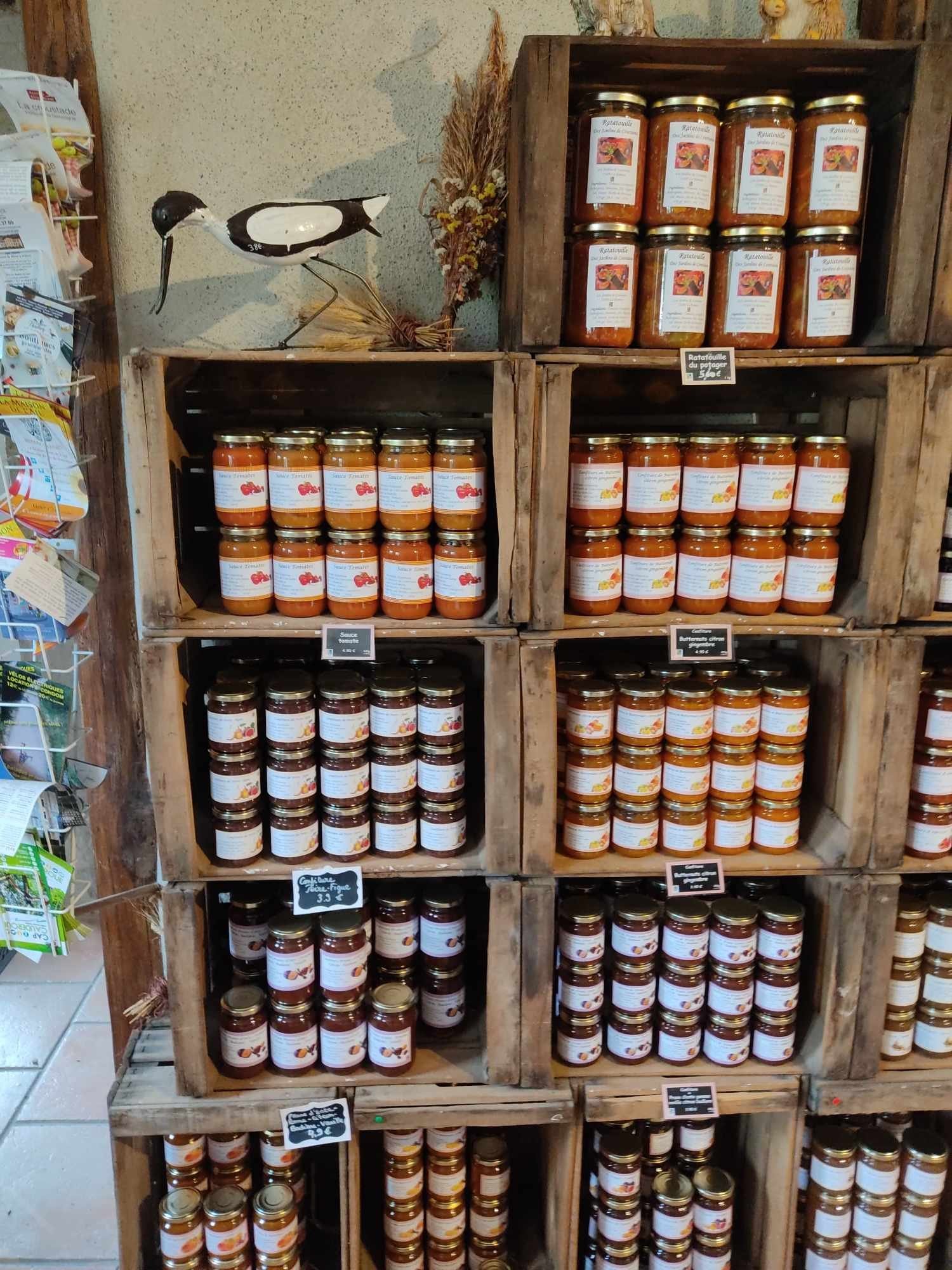 Des caisses en bois empilées servent d'étagères et présentent de nombreux pots de ratatouille, de sauce tomate et de confitures, avec un oiseau décoratif en haut à gauche.
