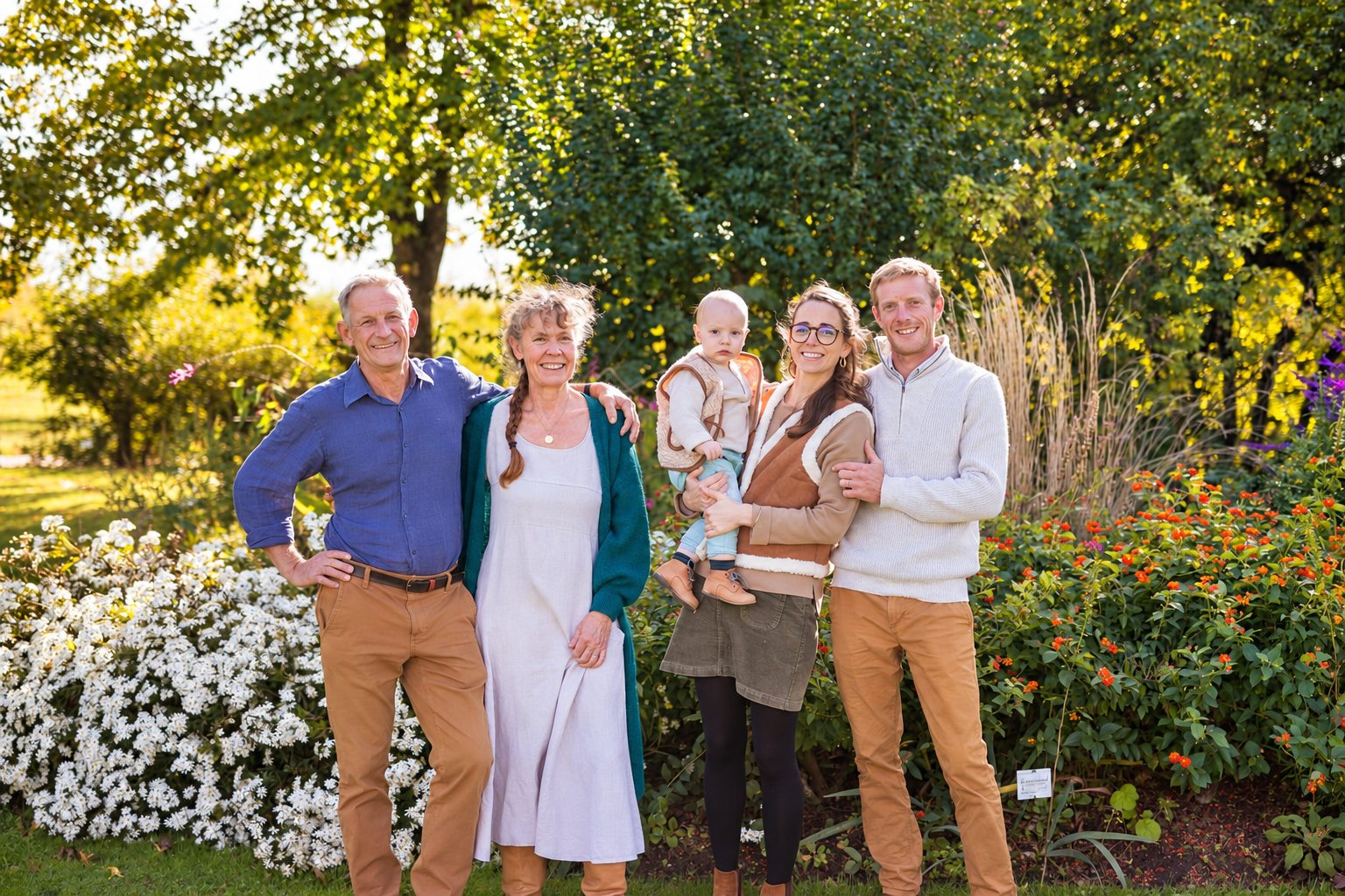 Quatre adultes souriants et un bébé posent côte à côte dans un jardin fleuri baigné de lumière dorée.