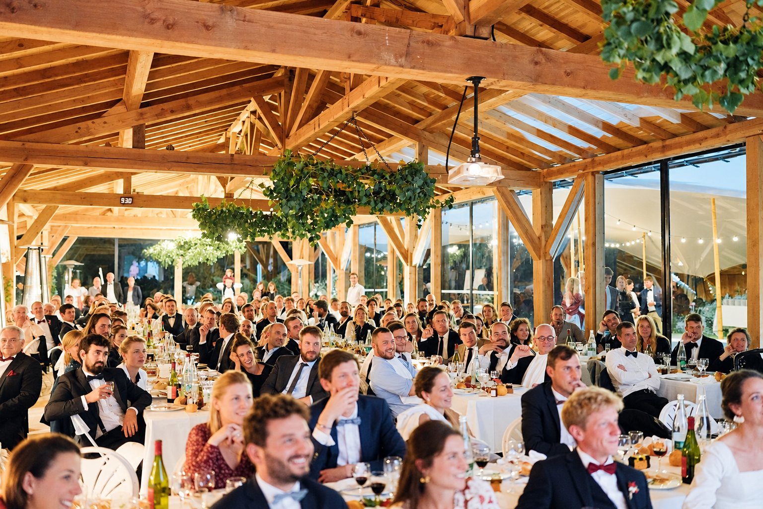 Une foule d'invités en tenue de soirée, assis à de longues tables, regarde en souriant dans une grande salle à charpente en bois décorée de feuillage suspendu.