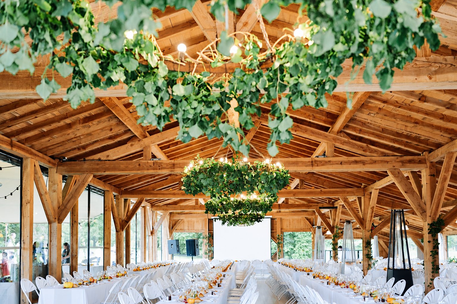 Une grande salle à charpente en bois, ornée de couronnes de feuillage suspendues, avec de longues tables blanches dressées pour un banquet.